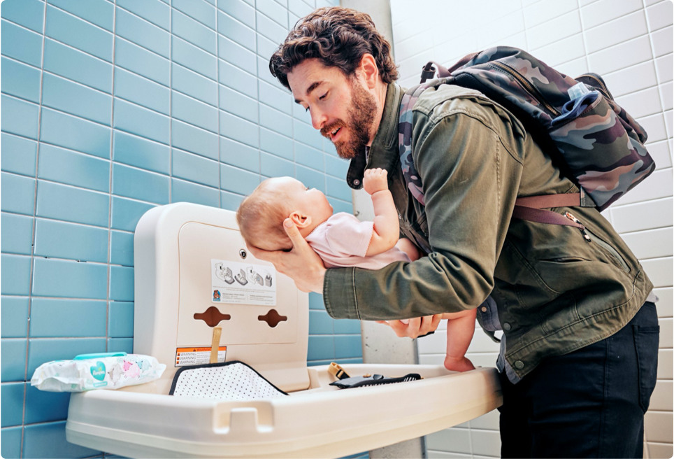 Dad holds baby over a changing table in a public restroom, with Pampers wipes on the side ready for use.