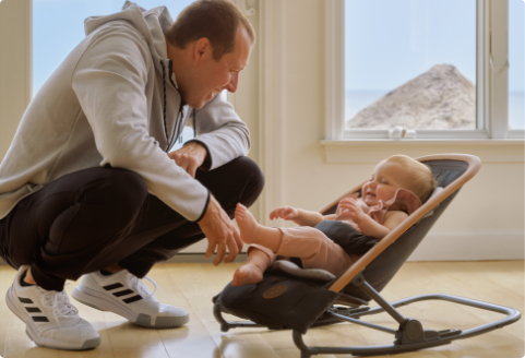 Dad crouches down to smile at baby seated in the Maxi Cosi Kori rocker.